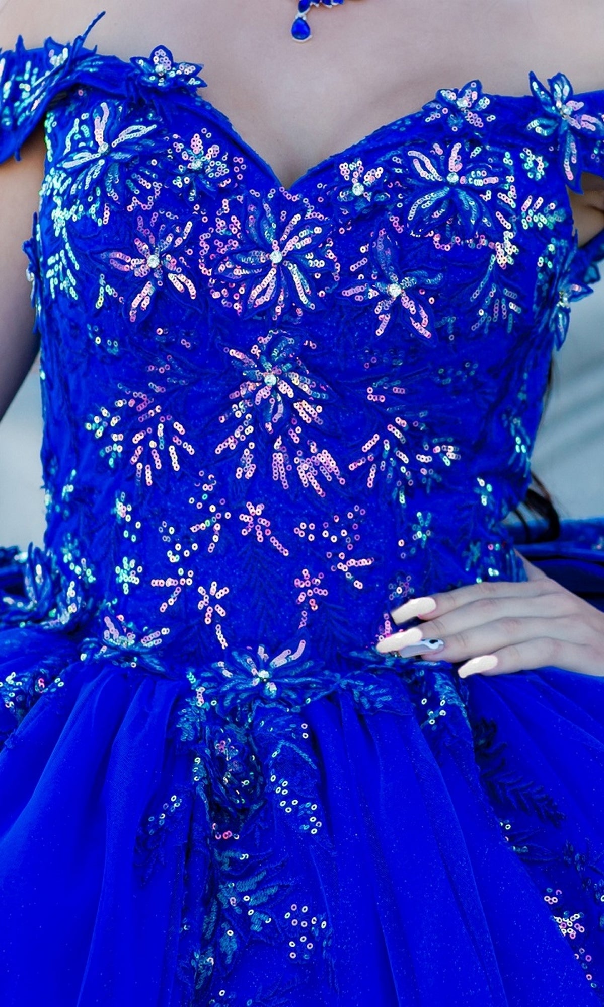 Someone wearing the Quinceanera Ball Gown 8100J, a bright blue off-the-shoulder dress featuring sequined floral patterns, iridescent beading, and a tulle skirt.